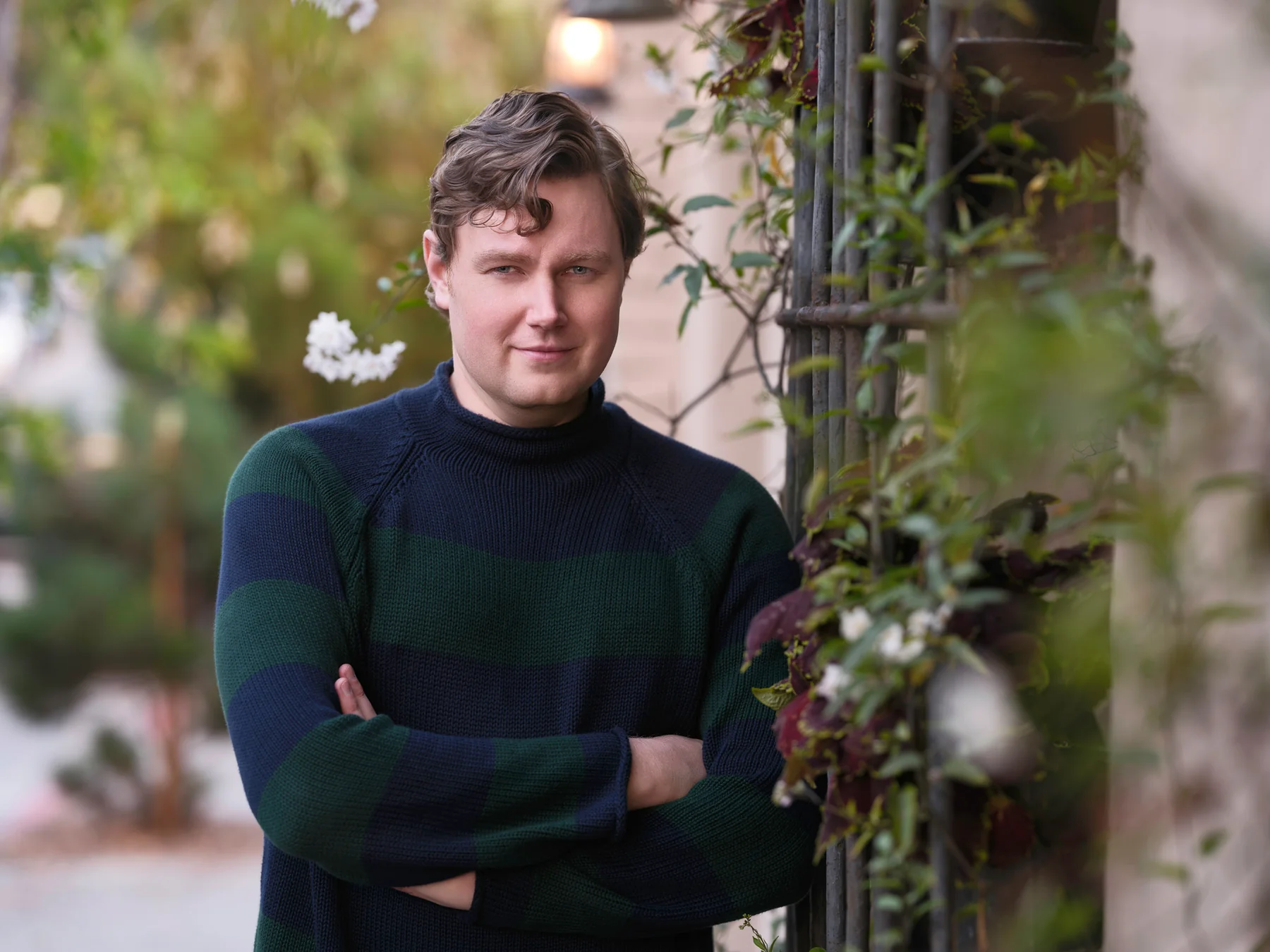 Brian Evergreen with arms crossed beside a vine-covered iron gate with white flowers in Carmel-by-the-Sea