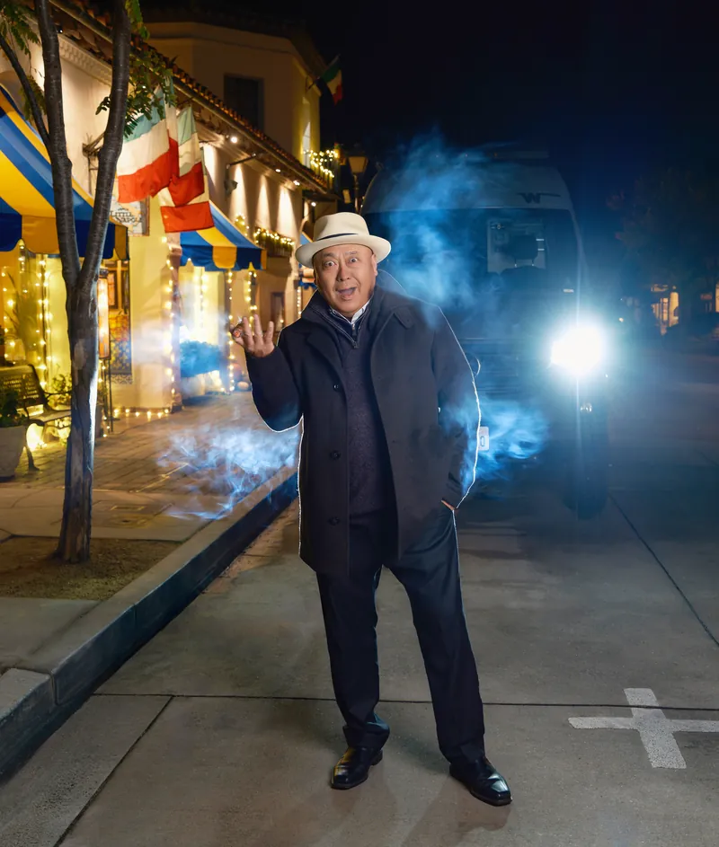 Man in a fedora and dark coat on a Carmel street at night with blue light and smoke behind him, editorial portrait by Manny Espinoza