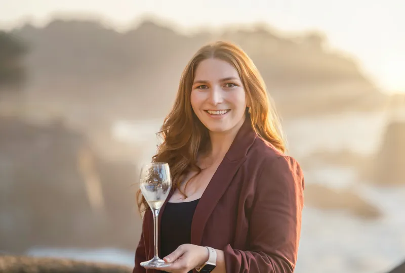 Woman in a burgundy blazer holding a champagne glass on the rocky Carmel coast at sunset, personal brand portrait by Manny Espinoza
