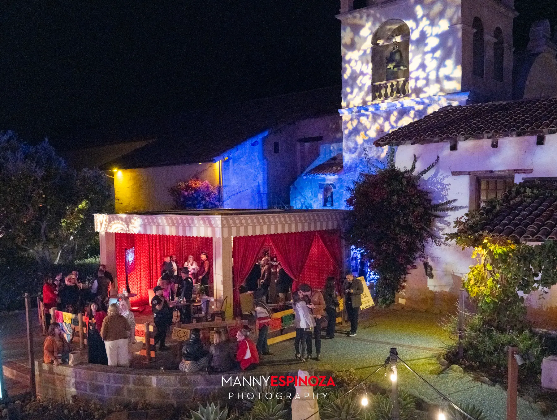 The Carmel Mission bell tower lit with patterned light above a red-draped tent and guests at Taste of Carmel 2026