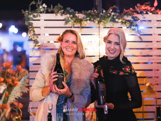 Two women holding wine bottles in front of a white wood backdrop with greenery at Taste of Carmel 2026