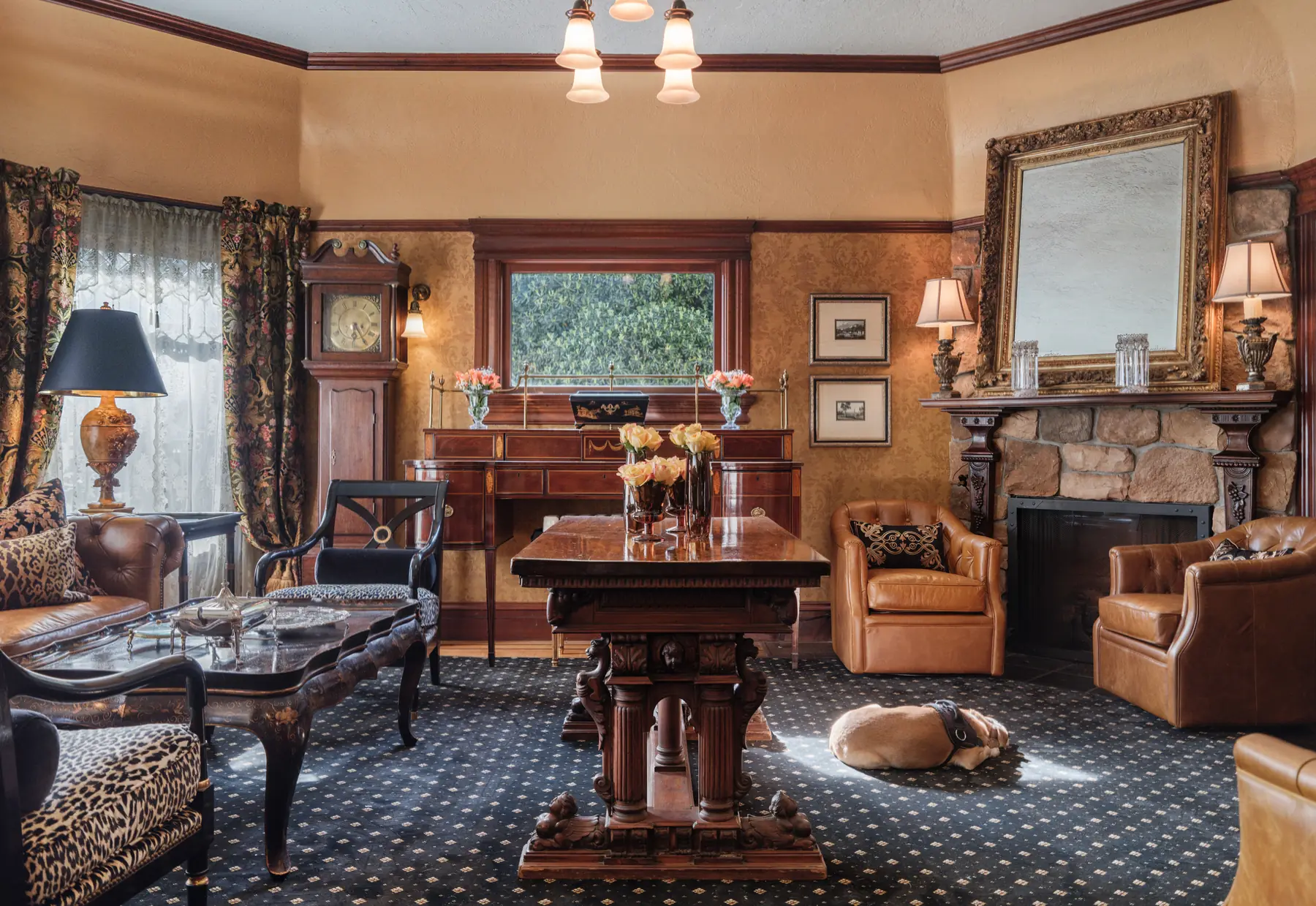 Living room at The Charles Napa Valley with a stone fireplace, antique grandfather clock, leather chairs and a dog resting by the hearth