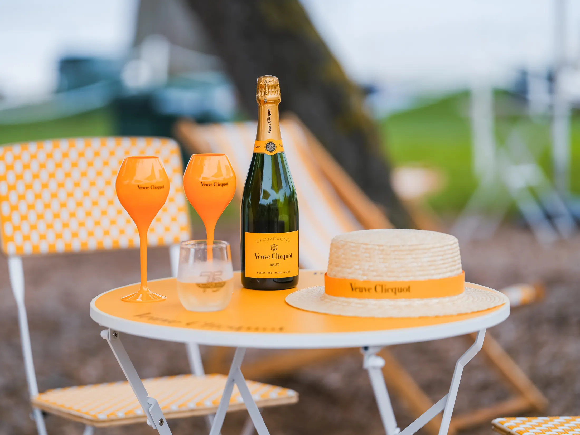 Veuve Clicquot champagne bottle, orange glasses, and branded straw hat on a bistro table at the Pebble Beach Concours d'Elegance