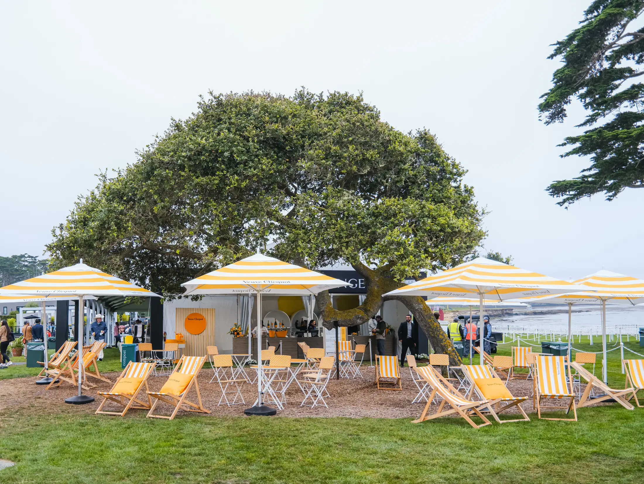 Veuve Clicquot lounge with striped umbrellas and deck chairs under an oak tree beside the 18th green at the Pebble Beach Concours d'Elegance