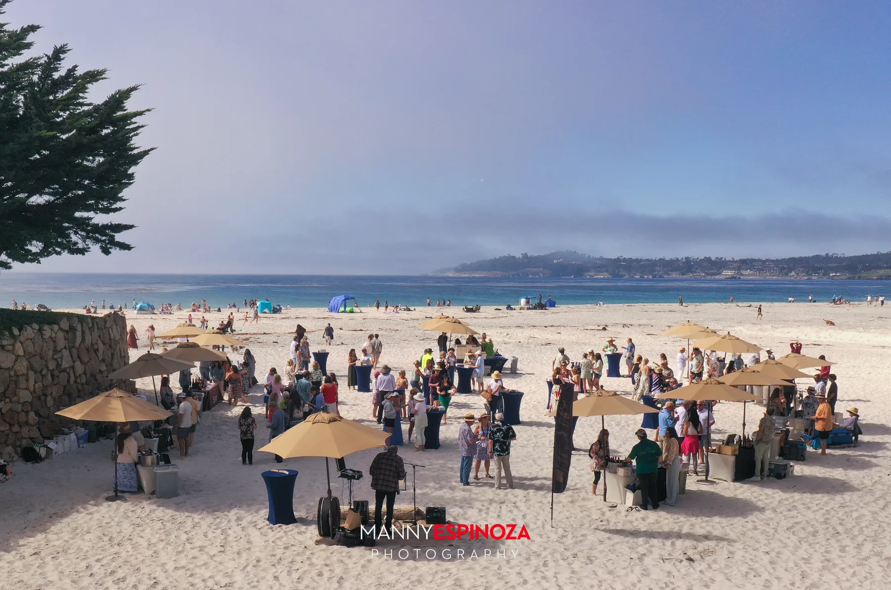 Drone view of the Walk for Freedom 2026 reception on Carmel Beach, with tan umbrellas and guests on the sand and Carmel Bay and Pebble Beach beyond