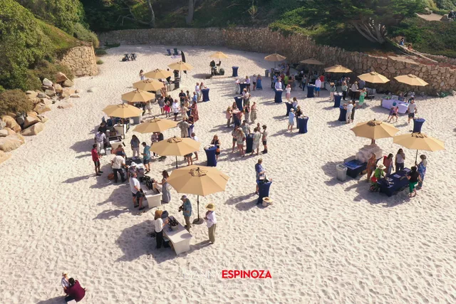 Drone view of the reception: tan umbrellas, blue cocktail tables and guests on the white sand below the stone seawall at Walk for Freedom 2026 on Carmel Beach
