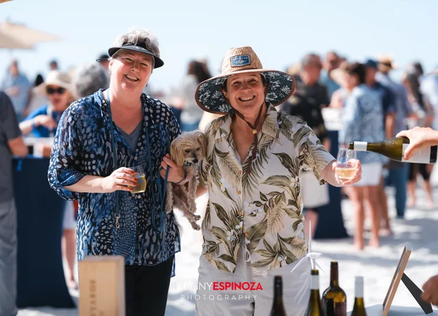 Two guests in straw hats toasting at a pouring table at Walk for Freedom 2026 on Carmel Beach