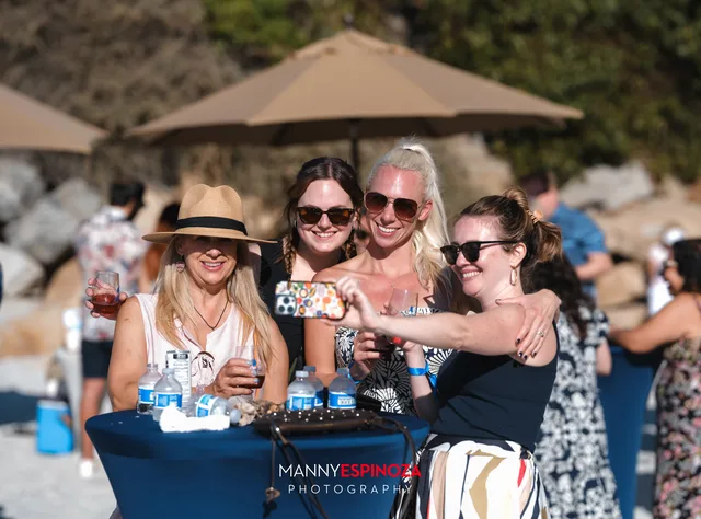 Four friends in sun hats laughing under an umbrella at Walk for Freedom 2026 on Carmel Beach