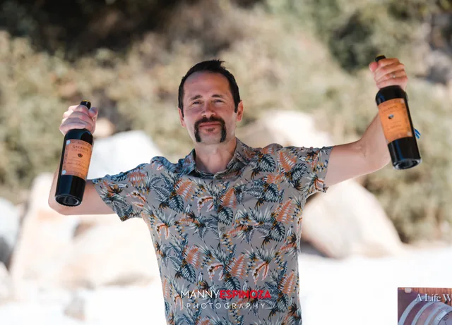 A man in a floral shirt grinning with a bottle held out in each hand at Walk for Freedom 2026 on Carmel Beach
