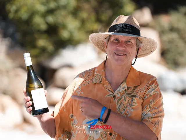 A winemaker in a straw hat and orange shirt holding a bottle of white wine at Walk for Freedom 2026 on Carmel Beach