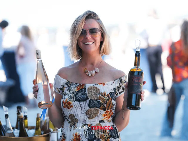 A woman in a floral off-the-shoulder dress holding two wine bottles at a pouring table at Walk for Freedom 2026 on Carmel Beach
