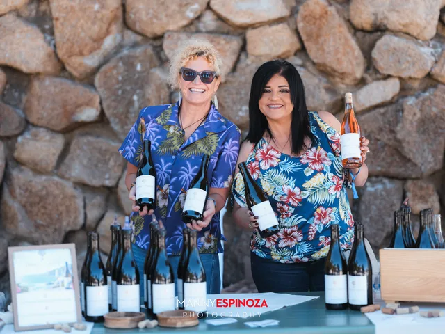 Two women in Hawaiian shirts holding bottles behind a lineup of wines below the stone seawall at Walk for Freedom 2026 on Carmel Beach